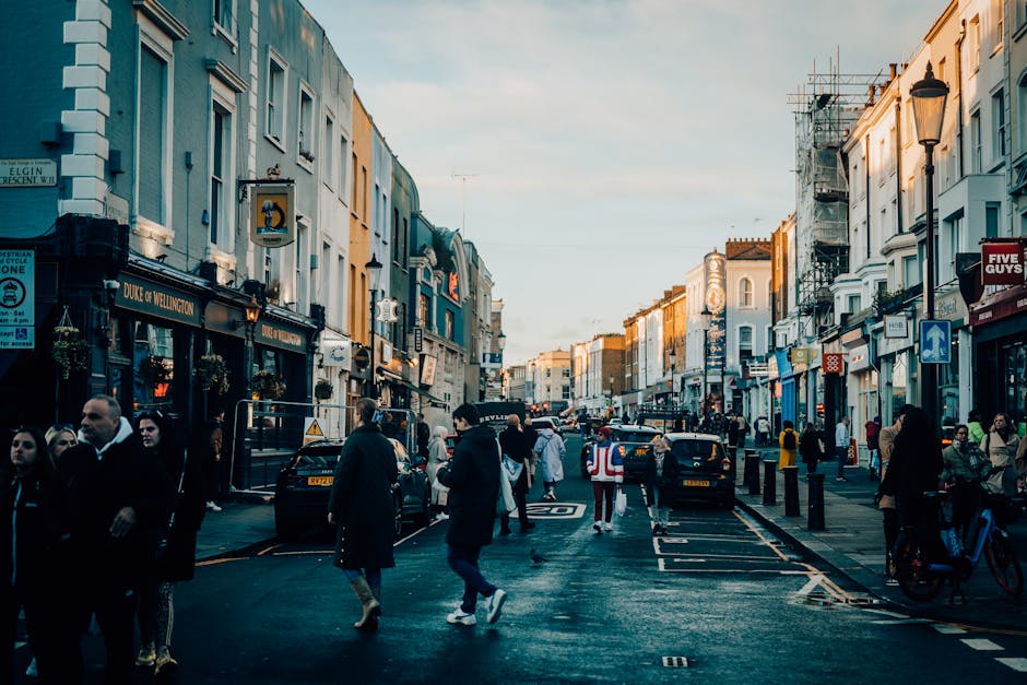 A busy urban street scene in a city with a mix of pedestrians walking along the pavement and crossing the road, and vehicles including parked cars and moving traffic on a wide, asphalt street. The buildings lining both sides of the street are multi-storey, with facades in various pastel and neutral tones, and some storefronts displaying signage such as 'Duke of Wellington' and others indicating retail shops, cafes, and businesses. Scaffolding is visible on one building on the right, suggesting ongoing maintenance or renovation. Street furniture includes black bollards, lampposts, and a sign indicating a one-way road. The lighting suggests late afternoon or early evening, with a clear sky casting a warm glow over the scene. The overall environment appears lively and active, typical of an area where private waste collection services, like those from Waste Disposal Notting Hill, might operate to manage commercial or residential rubbish removal in an alternative to municipal collection.