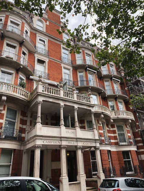 A multi-story Victorian-style residential building with a prominent white stone facade and ornate architectural details, including decorative columns and balustrades. The building features red brickwork with white accents around the windows and balconies, which have black wrought-iron railings. A central entrance with classical columns supports a small, curved balcony above, inscribed with the address ‘13-36’ and the name ‘Campden Hill Court.’ The building is partially obscured by green leafy tree branches in the foreground, framing the upper portion of the structure. Parked cars are visible along the street at the base of the building. This scene illustrates a typical example of private residential architecture in an urban area, aligning with the context of independent waste collection and alternative rubbish handling services often required for such distinctive properties, as provided by Waste Disposal Notting Hill.