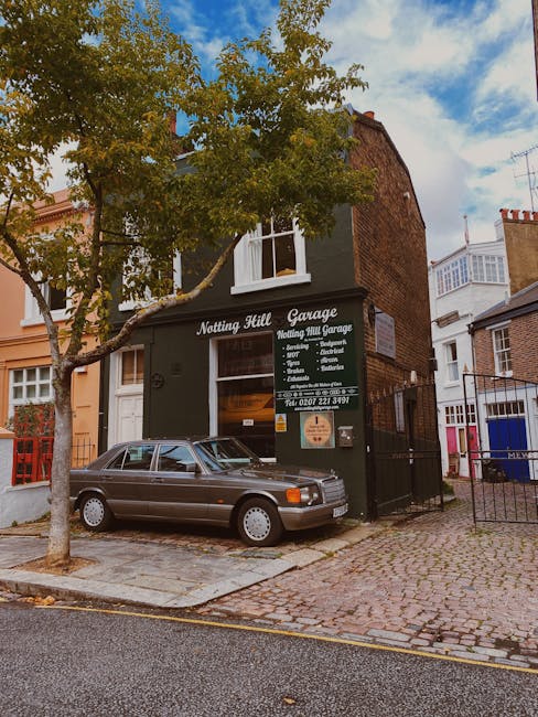 The image shows the facade of a small, dark green brick building housing Notting Hill Garage, with a large window above the entrance displaying signage for vehicle services. A vintage grey car with a smooth, matte finish is parked directly in front of the garage on a cobbled driveway. To the left of the building, there is a tree with green and yellow leaves, indicating a season of transition, its branches extending across the scene. The surrounding area includes other residential buildings with brick and stucco exteriors, some with bay windows, positioned along a narrow street. The sky above is partly cloudy with patches of blue, and soft daylight illuminates the scene evenly. The overall setting suggests an urban environment where private rubbish collection or on-site clearance services may be utilised, consistent with waste removal activities conducted by local or independent providers such as Waste Disposal Notting Hill.