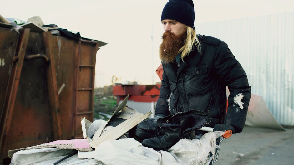 A multi-story Victorian-style residential building with a prominent white stone facade and ornate architectural details, including decorative columns and balustrades. The building features red brickwork with white accents around the windows and balconies, which have black wrought-iron railings. A central entrance with classical columns supports a small, curved balcony above, inscribed with the address ‘13-36’ and the name ‘Campden Hill Court.’ The building is partially obscured by green leafy tree branches in the foreground, framing the upper portion of the structure. Parked cars are visible along the street at the base of the building. This scene illustrates a typical example of private residential architecture in an urban area, aligning with the context of independent waste collection and alternative rubbish handling services often required for such distinctive properties, as provided by Waste Disposal Notting Hill.
