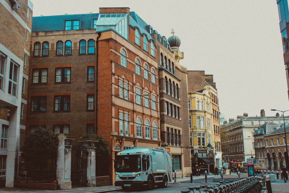 The image depicts a city street scene with a row of historic multi-storey buildings featuring a mix of brick and ornate finishes, typical of urban architecture in Notting Hill. In the foreground, a light grey waste collection vehicle from Waste Disposal Notting Hill is parked, positioned on the cobblestone street beside a sidewalk. The vehicle has a modern, smooth surface with visible windows and a compact, functional design suitable for urban rubbish collection. Surrounding the vehicle are several black metal bollards aligned along the curb, separating the pedestrian walkway from the roadway, with a bicycle rack filled with parked bikes visible on the right. The street is lined with a variety of buildings, including a cream-colored structure with large bay windows and decorative elements, and another with a rounded turret topped by a spherical dome. Natural daylight illuminates the scene through an overcast sky, casting soft shadows and highlighting the textures of the brick and stone façades. The scene reflects an urban environment where private waste collection services can operate amidst busy city life, supporting alternative rubbish handling options in densely built areas.