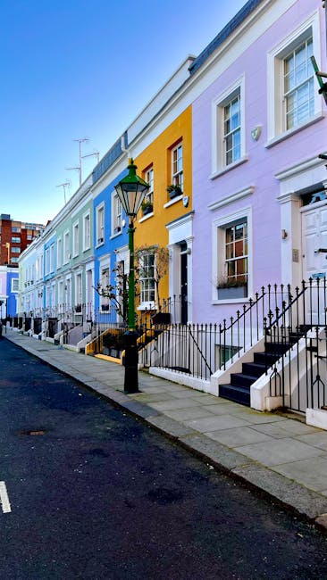A row of colourful terraced houses in Notting Hill, featuring pastel shades of blue, yellow, and lavender. The buildings have white-framed sash windows, some with window boxes, and black metal railings along the small front steps. A traditional black lamppost stands on the pavement in front of the houses, with a green decorative top. The street is clean, with a dark asphalt surface and a grey stone sidewalk, and the scene is lit with natural daylight, creating a clear and vibrant atmosphere. The overall setting reflects a typical residential area that could be associated with private waste collection or on-site clearance services, such as those offered by Waste Disposal Notting Hill for rubbish removal tasks at properties in this neighbourhood.