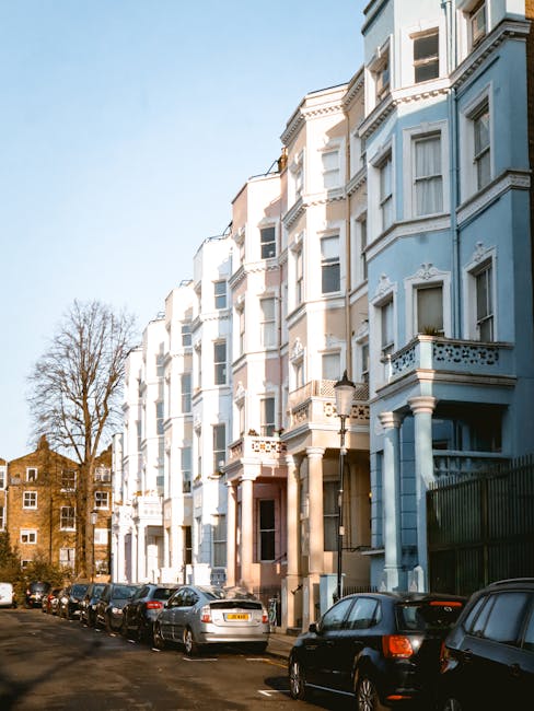 Eclectic row of multi-storey residential buildings painted in pastel shades of light blue and beige, featuring bay windows, ornate cornices, and decorative balcony railings. The buildings are situated along a narrow street with parked cars of various makes and models lining both sides, occupying the curb space. The foreground shows a tidy pavement with the vehicles neatly arranged, while behind the row of buildings, a leafless tree is visible against a clear blue sky, indicating a winter or early spring setting. To the left, a smaller brown brick building peeks into view, contrasting with the larger, more ornate structures. The scene suggests an urban environment with well-maintained architecture. The perspective emphasizes the height and details of the residential facades, which are characteristic of period architecture common in city boroughs that utilize private or alternative waste collection services, aligning subtly with the theme of independent rubbish removal solutions offered by Waste Disposal Notting Hill.