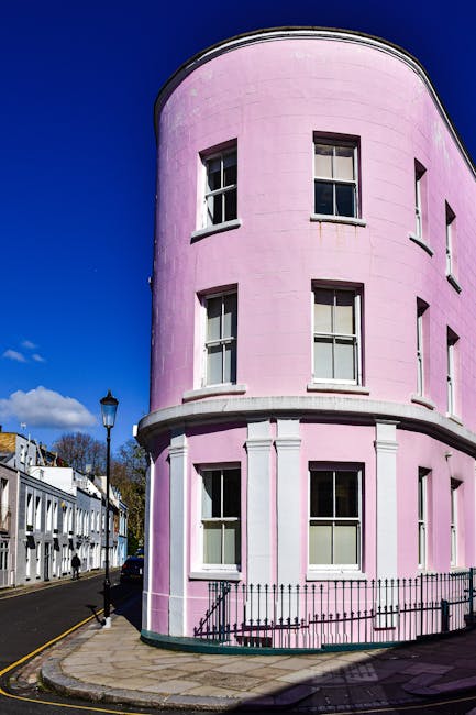 A row of colourful terraced houses in Notting Hill, featuring pastel shades of blue, yellow, and lavender. The buildings have white-framed sash windows, some with window boxes, and black metal railings along the small front steps. A traditional black lamppost stands on the pavement in front of the houses, with a green decorative top. The street is clean, with a dark asphalt surface and a grey stone sidewalk, and the scene is lit with natural daylight, creating a clear and vibrant atmosphere. The overall setting reflects a typical residential area that could be associated with private waste collection or on-site clearance services, such as those offered by Waste Disposal Notting Hill for rubbish removal tasks at properties in this neighbourhood.