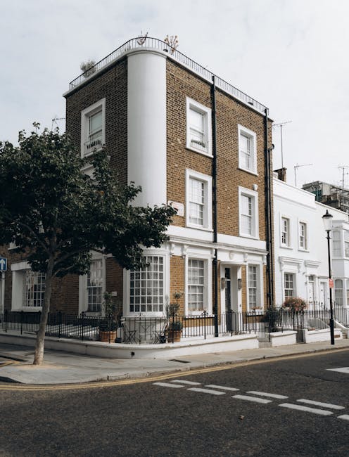 The image depicts a multi-story building situated at the corner of a street, featuring a combination of brickwork and white-painted facade elements. The building has three visible floors, with the upper floors clad in brown brick with white-framed sash windows, and a rounded corner section painted white. The ground floor appears to be a residential or commercial space with large windows framed by white trim, and a small black metal fence encloses a paved area with potted plants along the sidewalk. Next to the building, a small, leafy tree with dark green foliage partially obscures the lower windows on the left. The street in front is paved with asphalt, marked with crosswalk and lane lines, suggesting an urban environment emphasizing efficient vehicles and pedestrian movement. The scene is lit with natural daylight, indicating a clear or partly cloudy day. This setting could relate to private waste management or independent rubbish collection services operating within an urban residential area, in line with the theme of waste disposal and clearance services. The overall scene is neutral, factual, and focused on the urban environment with no visible rubbish or waste materials in view.