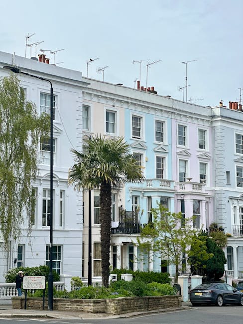 A row of three-storey Victorian-style terraced houses painted in light pastel shades, predominantly white with some subtle blue accents, situated along a street with a small front garden featuring a tall palm tree, shrubs, and small trees. The buildings have decorative architectural details such as cornices and window surrounds, with multiple sash windows on each floor, some of which are open or fitted with security bars. In the foreground, there is a black lamppost, a parked dark-colored car, and a wooden signboard indicating the residential area. The pavement appears clean and well-maintained, with a low brick wall enclosing the garden beds. The sky above is cloudy, affecting the overall soft lighting of the scene. This image, possibly captured during daylight hours, relates to the context of independent waste management and rubbish removal, reflecting a typical urban environment where private rubbish collection services might operate to complement or provide alternatives to municipal rubbish rules.