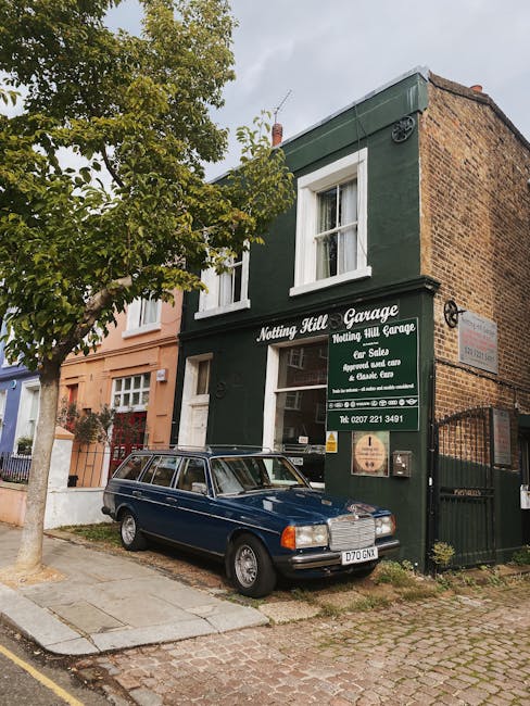 A two-storey building painted dark green with white window frames, housing the 'Notting Hill Garage' business, with a large green sign displaying services such as sale, serviced, and repair of vehicles. The building is attached to a brick wall on the right side, and a black metal gate is visible on the right edge. In front of the garage, there is a classic dark blue station wagon parked partly on the paved sidewalk and partly on the cobblestone street. To the left of the vehicle, a tree with green foliage extends branches overhead, partially obscuring the upper part of the building. The scene is set on a quiet street with neighboring buildings painted in pastel shades, such as light pink and cream, and the pavement is bordered by a curb. The sky above is overcast, providing diffuse natural light, and the overall scene suggests an urban environment where private waste collection or rubbish removal services like Waste Disposal Notting Hill may support local property management or independent collection efforts, keeping the area clear of rubbish and debris.
