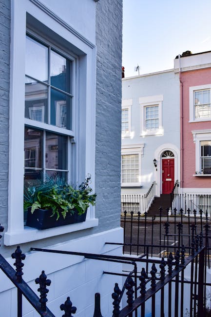 A two-storey brick building with a dark green facade, labeled 'Notting Hill Garage' and advertising services such as vehicle repairs and diagnostics, is situated on a cobblestone street. In front of the garage, a grey vintage sedan with a metallic finish is parked parallel to the building, partially shaded by a large deciduous tree with green leaves that extends over the sidewalk. The tree’s trunk is slim, with textured bark, and its branches spread outward, casting dappled light on the car and pavement. The surrounding area includes adjacent residential buildings with white and brick exteriors, some with bay windows, and metal railings enclosing small front yards. The sky above is partly cloudy with patches of blue visible, providing a neutral daylight setting typical for a street-level view associated with waste management and debris clearance services from private providers such as Waste Disposal Notting Hill, implied by the presence of the garage used for vehicle and equipment storage related to rubbish collection and site clearance activities.
