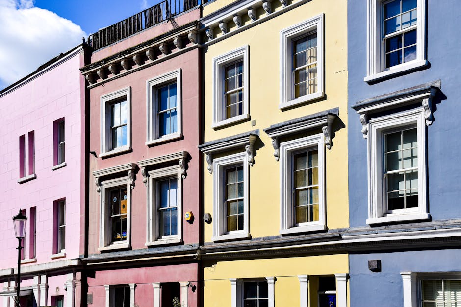 A row of three adjacent multi-story residential buildings painted in pastel shades of pink, yellow, and blue, with classic white-framed sash windows and decorative cornices. The pink building features a main entrance door with glass panels and a small mailbox beside it, while the yellow and blue buildings have similar window arrangements on each floor. The facades are smooth with a subtle texture, and the buildings are situated close together on a city street, with a black streetlamp visible on the left side of the image. The scene is illuminated by natural daylight, casting soft shadows on the buildings' surfaces, creating a vibrant yet neutral urban atmosphere that aligns with private property maintenance and the potential for external rubbish collection or on-site clearance services, as offered by Waste Disposal Notting Hill.