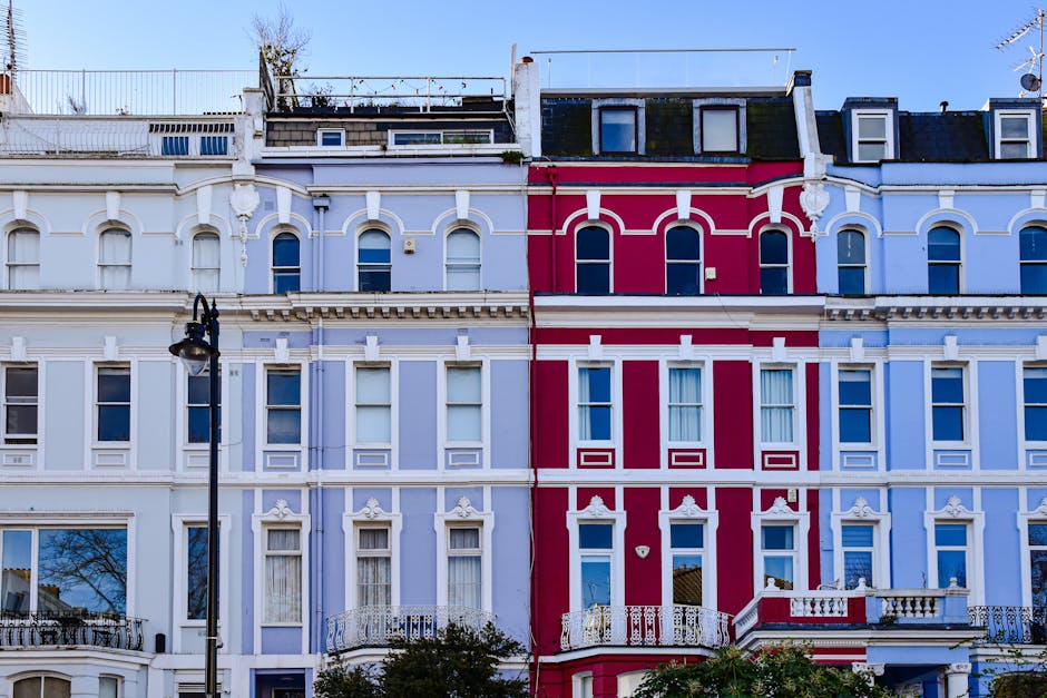 A row of white and pale blue Victorian-style residential buildings on a city street, with multiple bay windows and decorative molding, shows a typical London terrace. In the foreground, parked cars of various makes and models, including small hatchbacks and sedans, line both sides of the street. The pavement is partially visible, with some greenery and a gate on the right side, possibly leading to a garden or private entrance. To the left, leafless trees are seen against a clear, bright blue sky, suggesting a late autumn or winter season. The scene appears quiet and well-maintained, reflecting a typical urban residential area where private waste disposal or rubbish removal services from Waste Disposal Notting Hill might be relevant for managing household or building waste between local collection days.