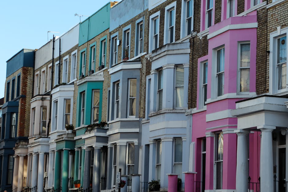 A row of Victorian-style terraced houses with bay windows and decorative cornices, painted in pastel shades including white, pink, blue, and teal. The buildings are constructed from red brick with some areas painted to enhance their appearance. Each house features small front gardens with iron railings, and some have potted plants or flower boxes on the window sills. The sky above is clear with a soft blue tone, and the sunlight casts gentle shadows on the facades, highlighting the architectural details. The image captures an urban residential street scene, typical of a densely built area, with an emphasis on the colorful, well-maintained exteriors of the houses. This scene reflects a typical UK street where private property maintenance and aesthetic enhancements are part of everyday urban life, aligning with waste disposal and rubbish management practices in residential environments supported by local service providers such as Waste Disposal Notting Hill.