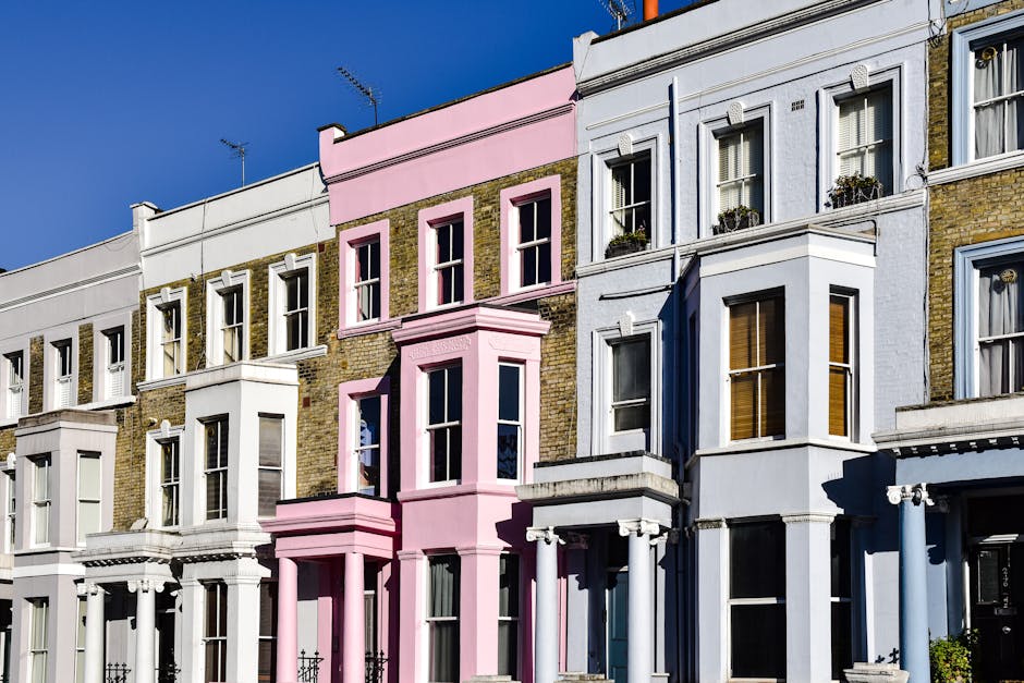 A row of multi-storey Victorian terraced houses with detailed architectural features, situated along a street with a clear blue sky overhead. The buildings have a mixture of painted brickwork in white, light blue, and pink, with some sections featuring exposed yellowish-brown brick. The central house is painted pink with a prominent bay window on the first and second floors, and decorative mouldings above the windows. To the left and right, the neighbouring houses are painted in soft pastel shades with white window frames, some with flower boxes on the windowsills. The facades include ornate cornices, brick columns, and decorative window trims, creating an elegant, historic appearance. The sunlight casts sharp shadows, highlighting the textures of the painted stucco, brick, and wooden window frames. While not shown in the image specifically related to rubbish removal, the setting suggests an urban residential area that might be serviced by independent waste disposal providers such as Waste Disposal Notting Hill for precise and private waste management arrangements.